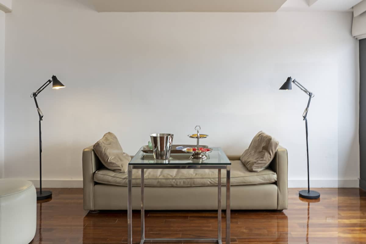 A modern living room with a glass-top coffee table, a beige sofa, and two floor lamps.