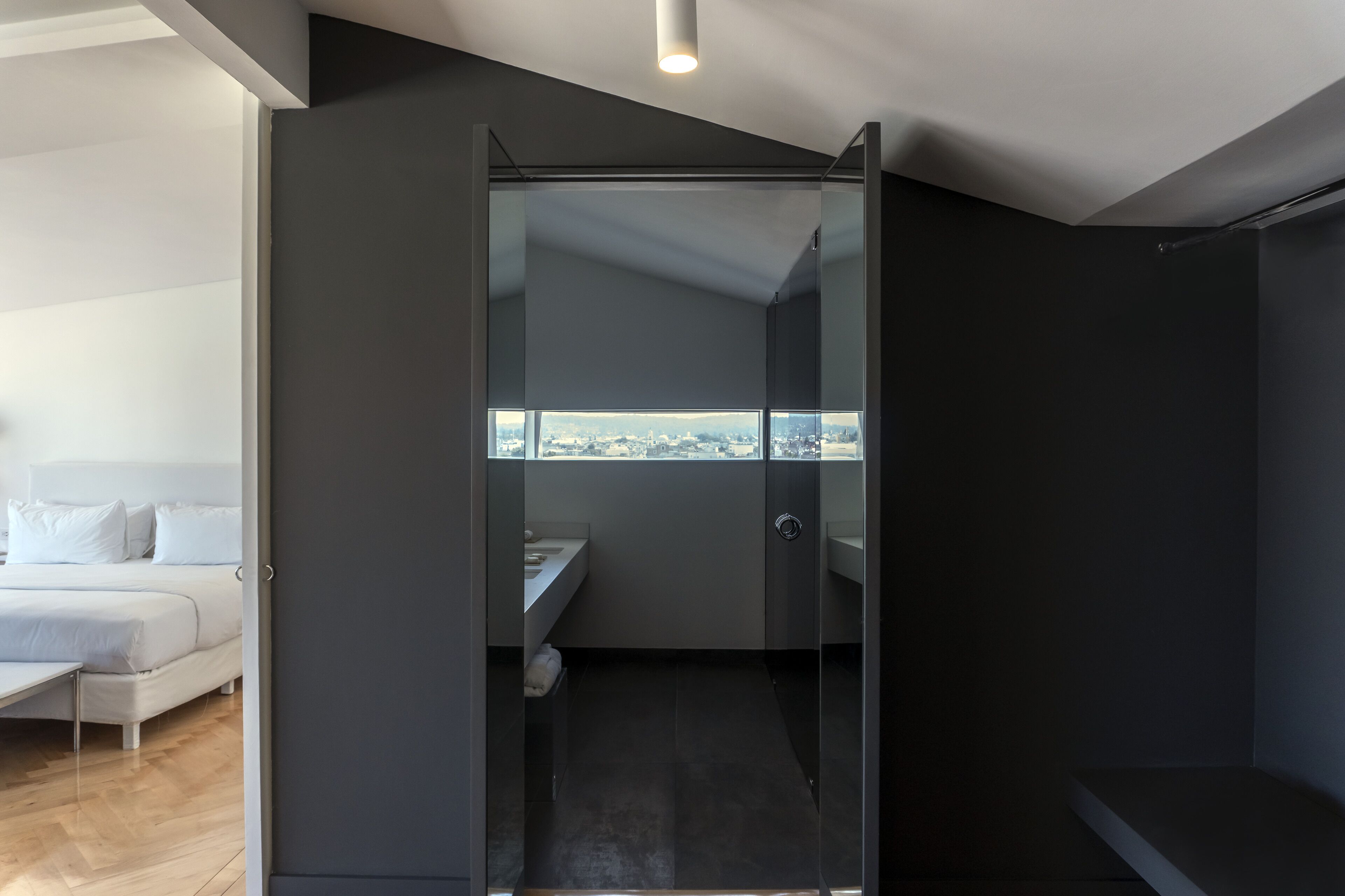 Una habitación de hotel moderna con ducha de vidrio, cama con ropa blanca y vista a la ciudad.