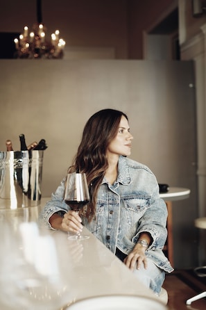 Una persona sentada en un bar, sosteniendo una copa de vino, con un recipiente metálico con cubiertos y un candelabro al fondo.