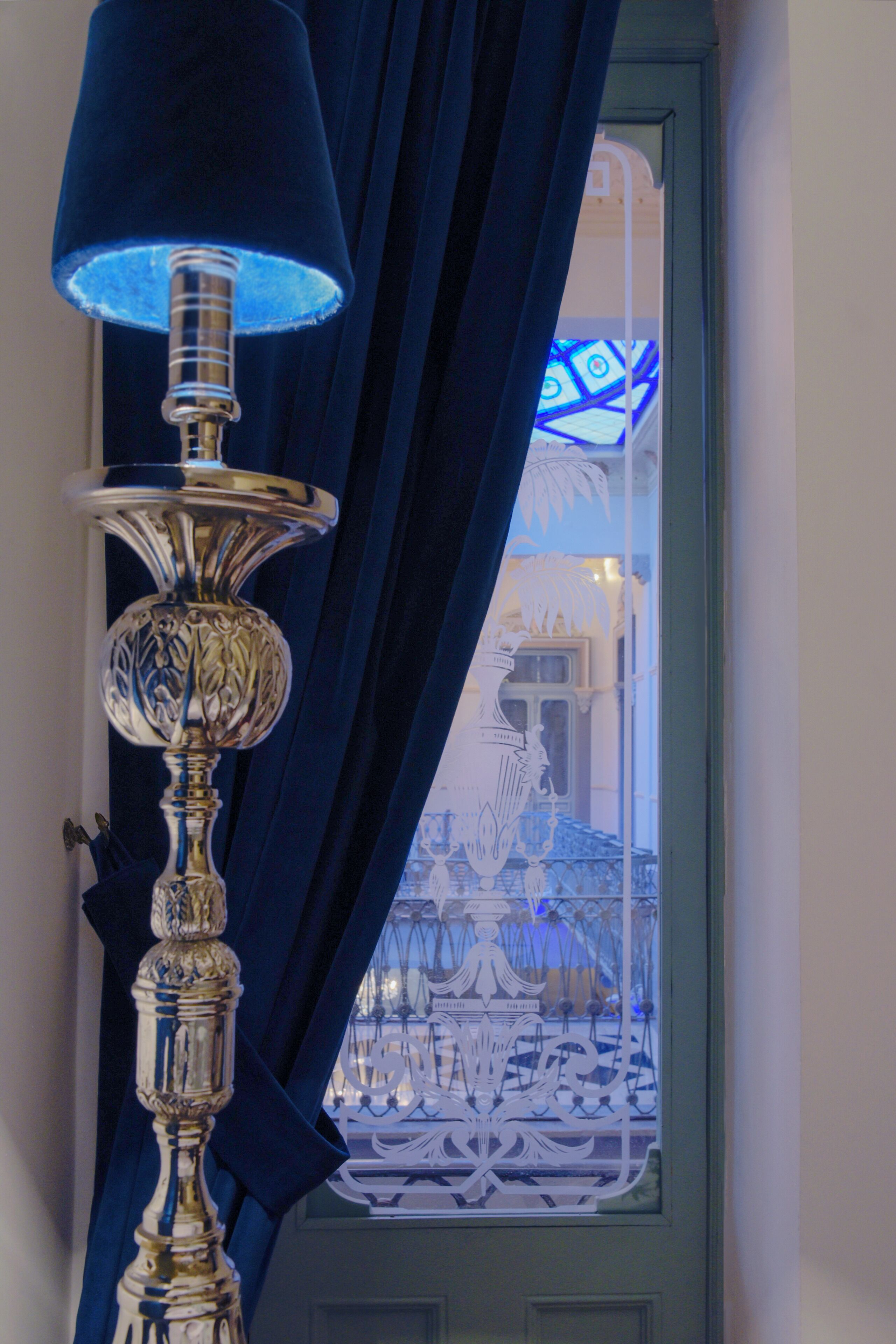 A lamp with a blue shade and a decorative base, positioned in front of a window with blue curtains.