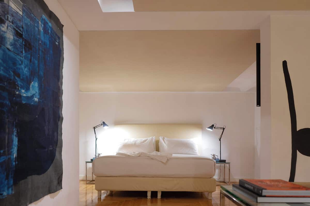 A bedroom with a large bed, two bedside tables, a nightstand, a lamp, and a partial view of a painting.
