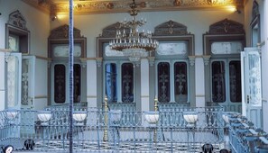 Una sala majestuosa con techo ornamentado y una araña, que presenta una larga mesa con sillas y una barandilla decorativa.