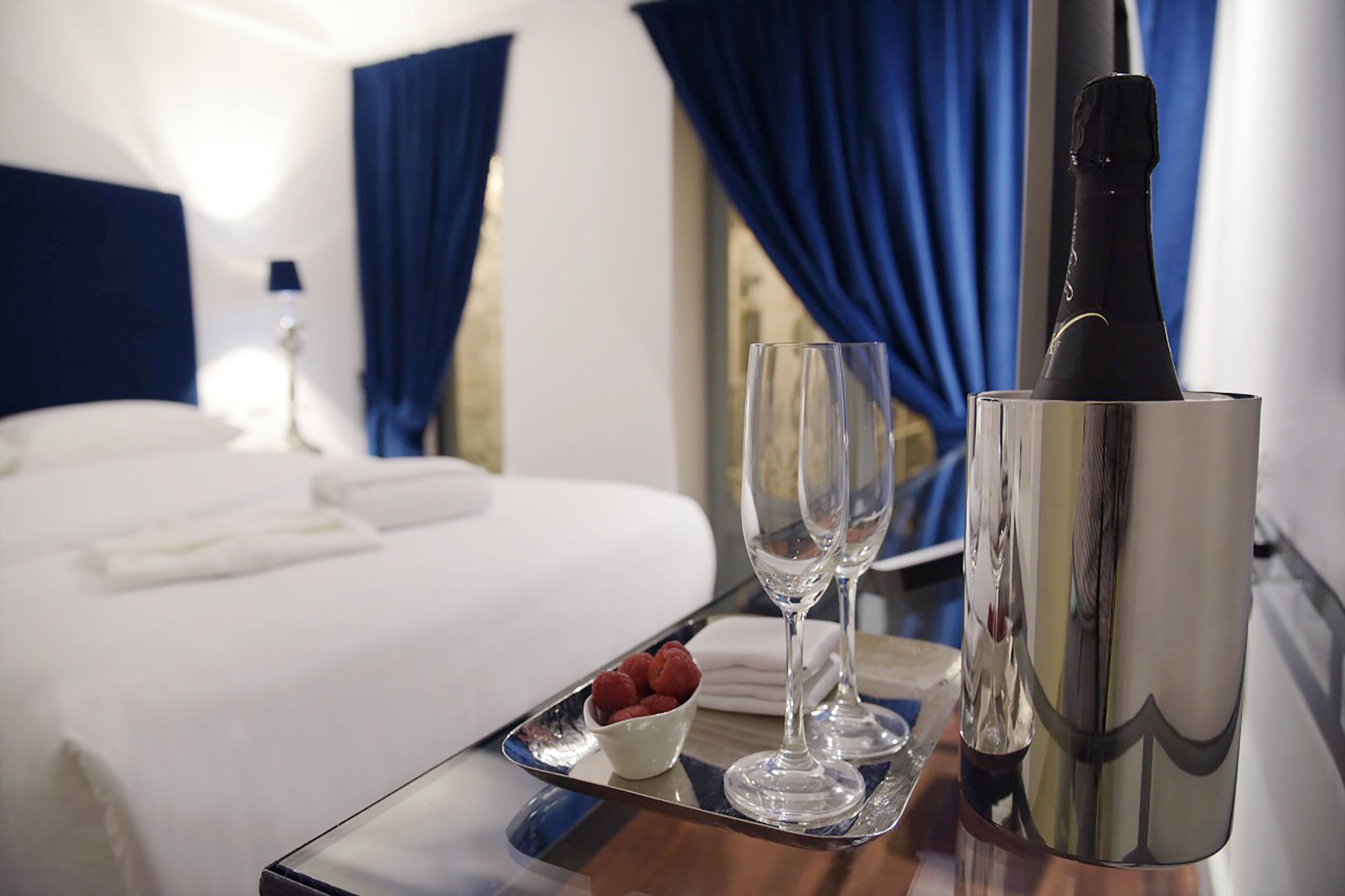 Ein Hotelzimmer mit einem ordentlich bezogenen Bett, einem Tablett mit einer Champagnerflasche, zwei Weingläsern und einer Schale Himbeeren.
