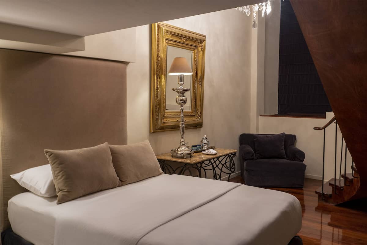 A hotel room with a large bed, a desk with a lamp, a chair, and a staircase.