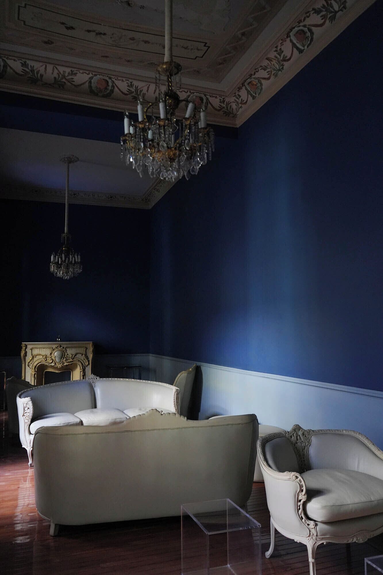 A room with a dark blue wall, a white sofa, and a white armchair. There is a chandelier hanging from the ceiling.