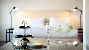 Uma mesa com um suporte para sobremesas, dois copos de coquetéis e um prato com petiscos em quarto de hotel, com uma cama e mesas de cabeceira ao fundo.
