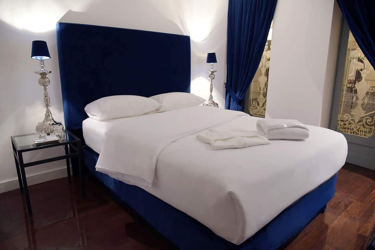 A neatly made bed with white linens and two pillows, flanked by two bedside tables with lamps, in a room with a blue headboard and a large window with blue curtains.