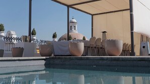 Uma área junto à piscina com um espaço coberto para sentar, mobiliário exterior e vista para um edifício com cúpula ao fundo.