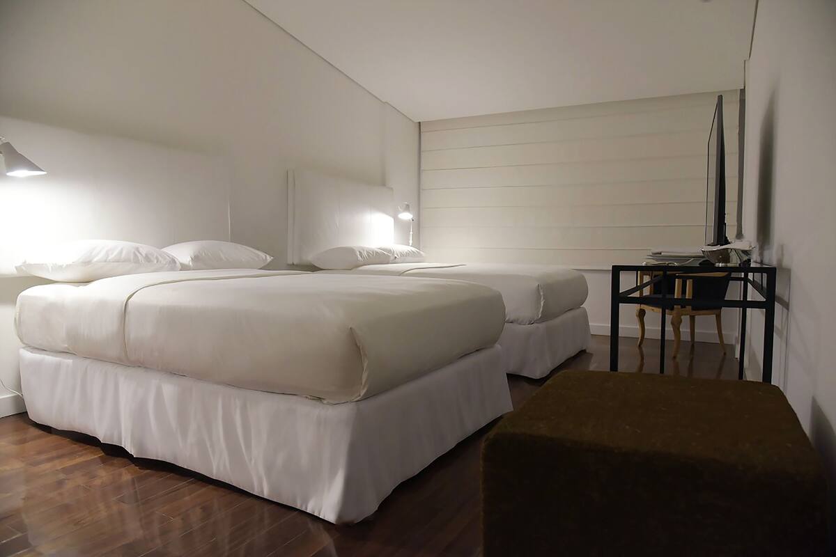 A bedroom with two beds, a nightstand, a desk, and a brown ottoman.