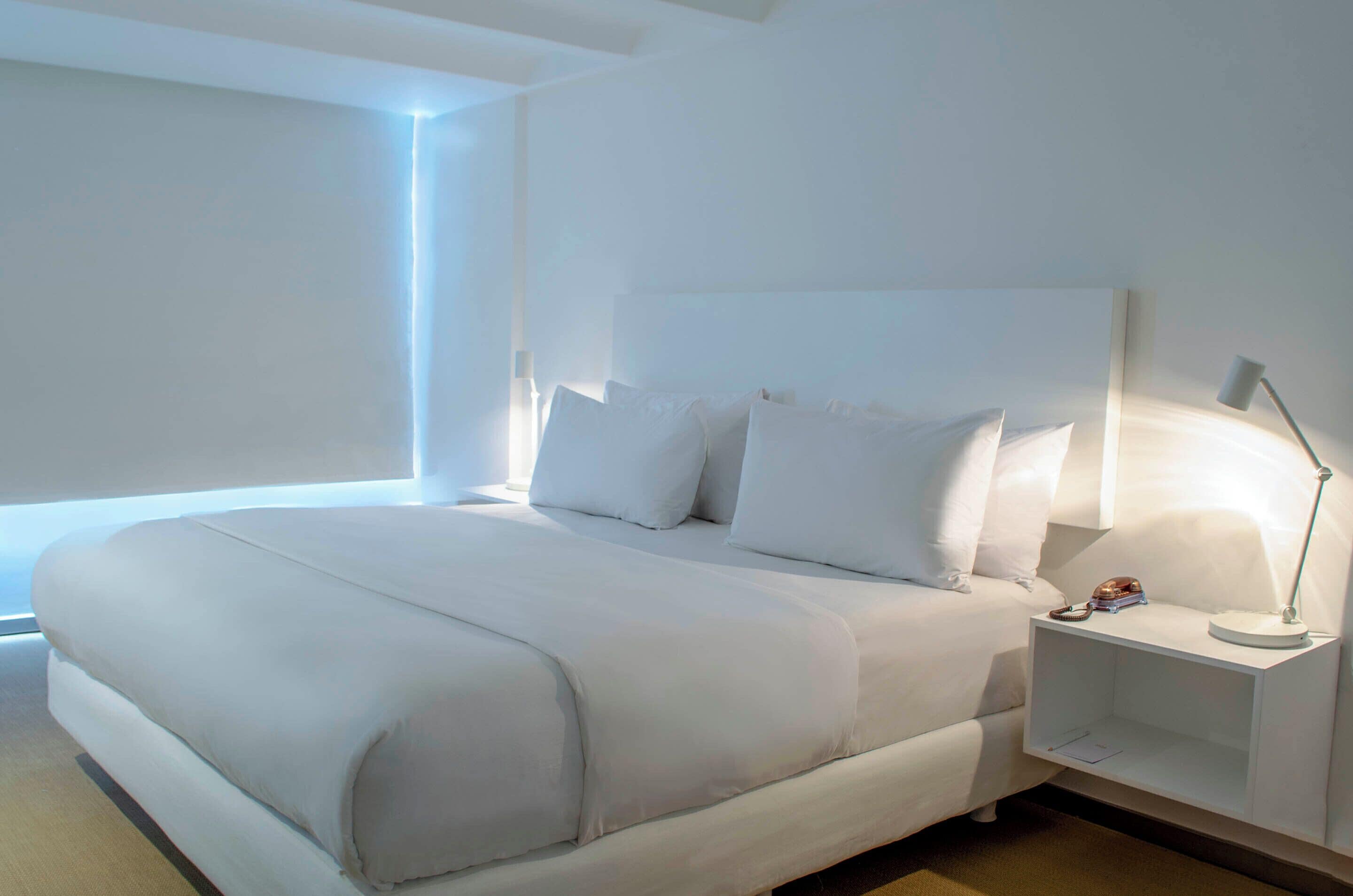 A modern bedroom with a large bed, white bedding, a bedside table with a lamp, and a nightstand with a phone and book.