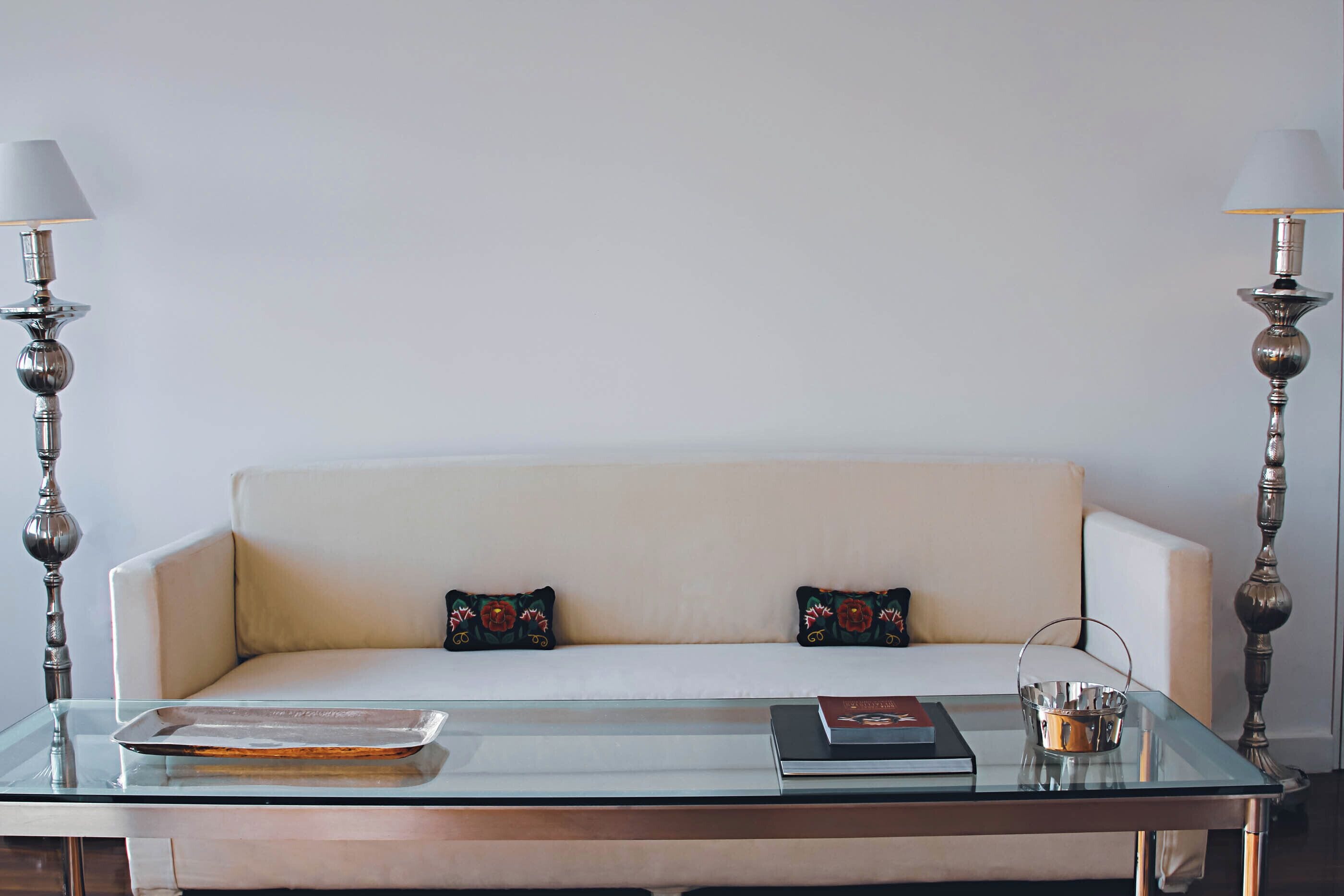Una sala de estar moderna con una mesa de centro de vidrio, un sofá blanco con cojines estampados y dos lámparas de pie decorativas.