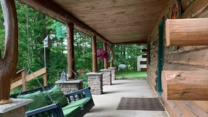 A covered porch with a swing seat, stone pillars, and a view of the surrounding forest. - Beautiful Log Cabin 11ac. 4 br. 2.5 ba. on snowmobile trails in Manistee forest. (Township of Branch)