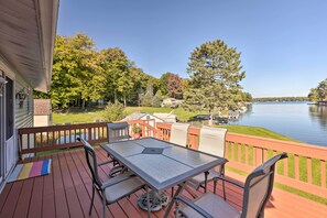 A wooden deck with a table and chairs, overlooking a lake with boats and a house in the background. - Lakefront Ludington Retreat w/ Kayaks & Fire Pit! (Ludington)