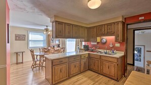 A kitchen with wooden cabinets, a central island, a dining area with chairs, and a window with blinds. - Lakefront Ludington Retreat w/ Kayaks & Fire Pit! (Ludington)