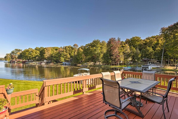 A wooden deck with a table and chairs overlooking a lake, surrounded by trees and boats. - Lakefront Ludington Retreat w/ Kayaks & Fire Pit! (Ludington)