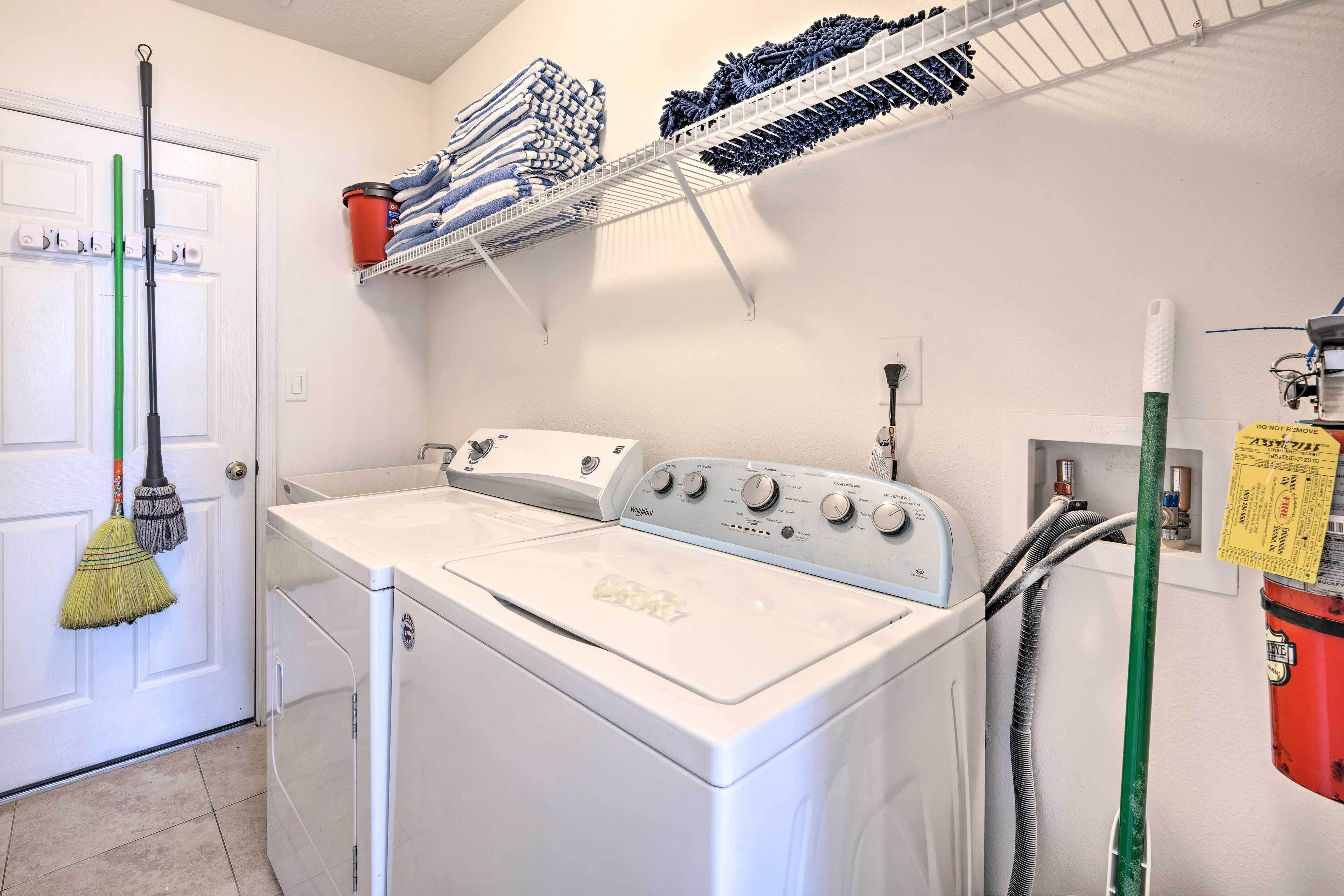 A laundry area with a front-loading washer and dryer, a broom, and a shelf with folded towels and cleaning supplies.