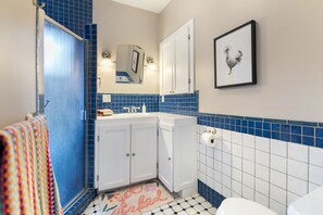 Casa de banho com lavatório branco, paredes azulejadas em azul e branco, um quadro com uma galinha e uma toalha colorida.