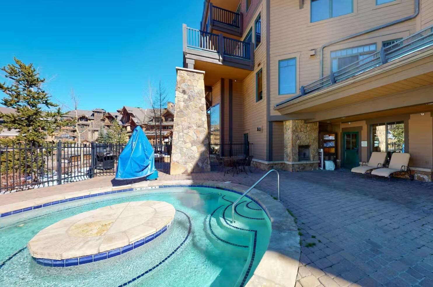 Une piscine bordée de pierres, un espace détente couvert et un bâtiment doté de balcons.
