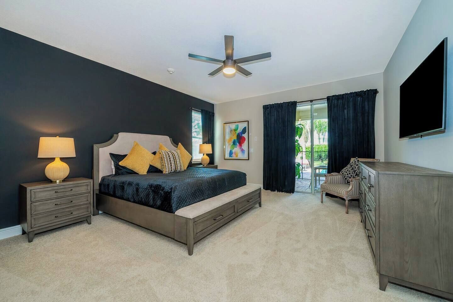 A bedroom with a bed, bedside tables, a dresser, a ceiling fan, and a view of the outdoors.