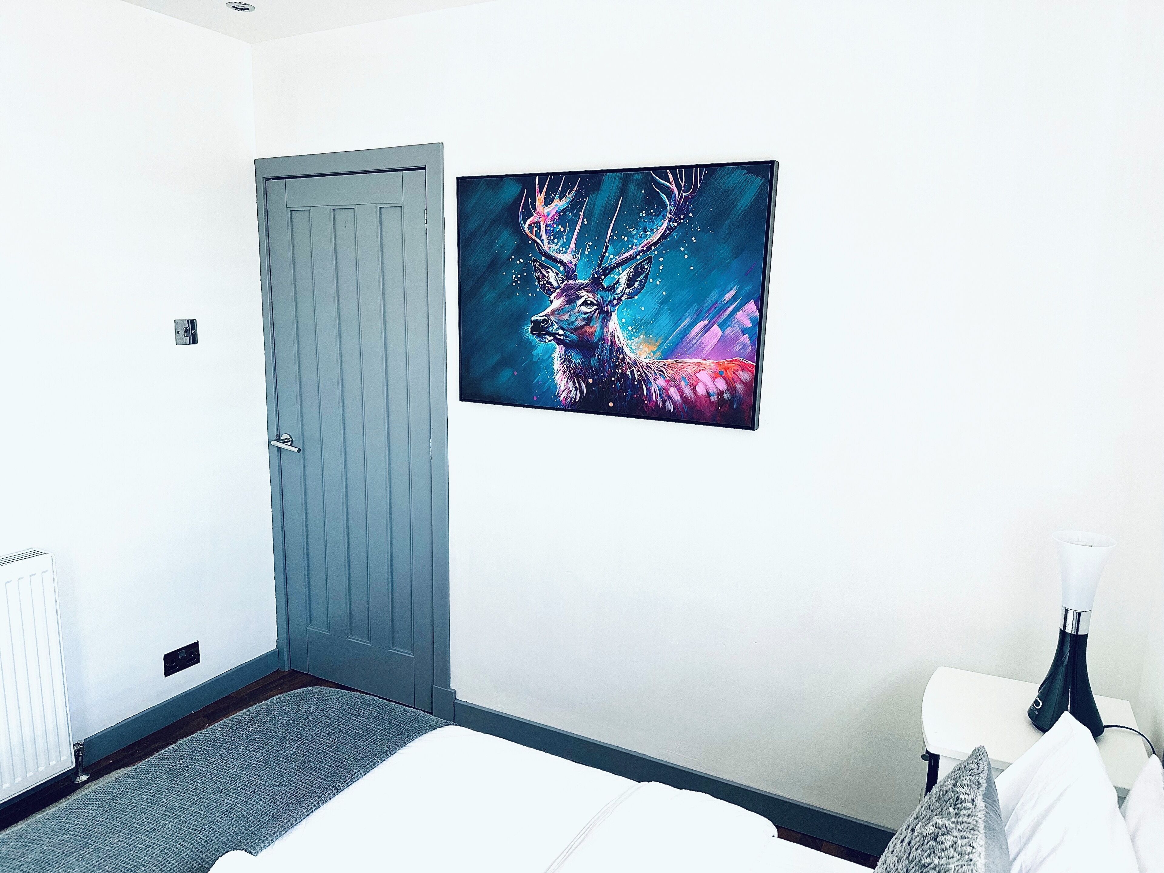 Quarto de hotel com porta, cama, mesa de cabeceira e uma obra de arte de um veado na parede.
