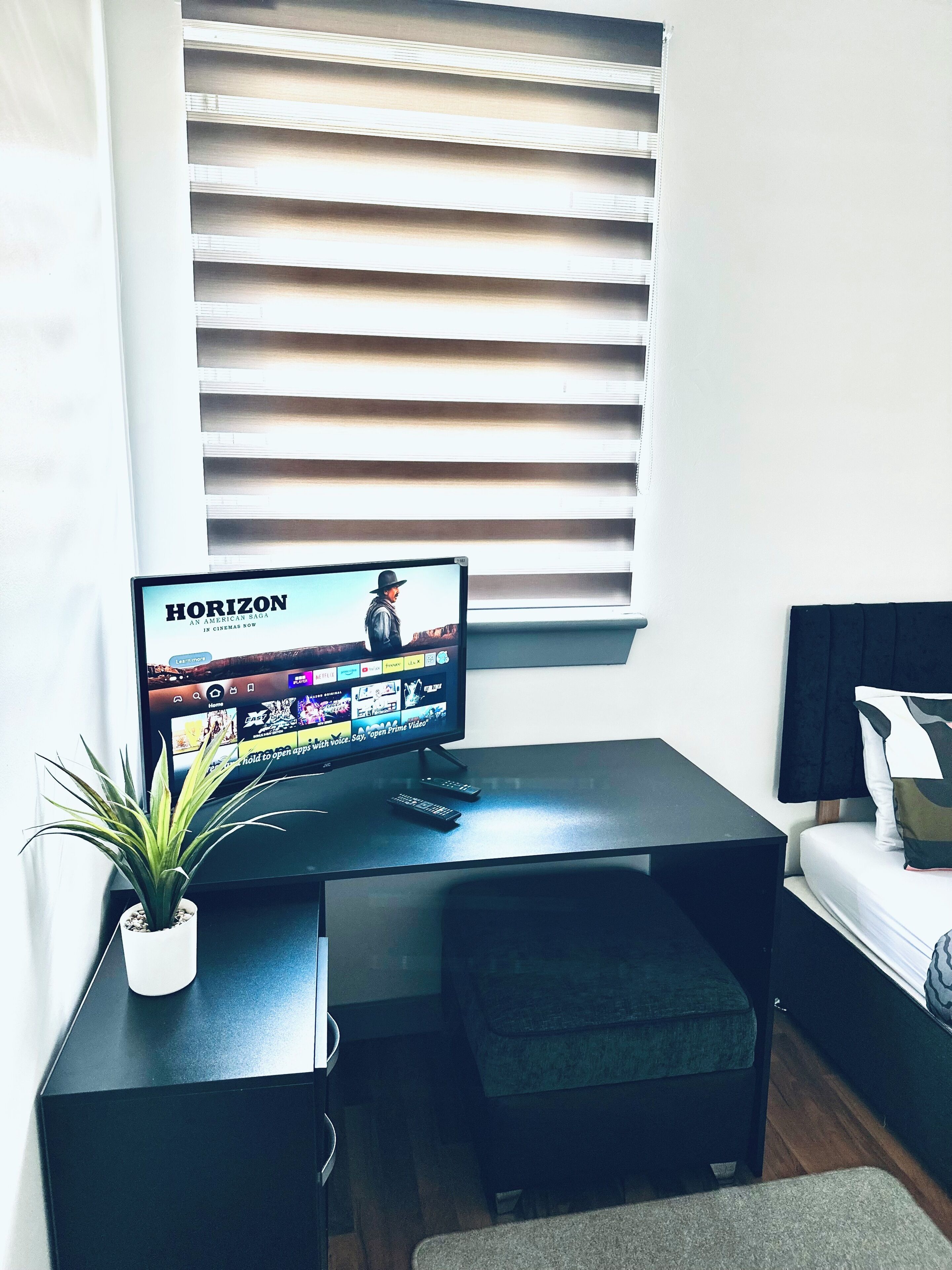 A modern room with a TV displaying 'Horizon' on the screen, a small desk with a plant, and a bed with a black headboard.