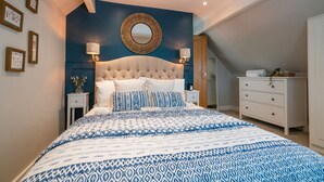 A bedroom with a tufted headboard, a large bed with patterned bedding, a round mirror, and a dresser. - Pilgrim Cottage - Luxury Cottages (Chester)