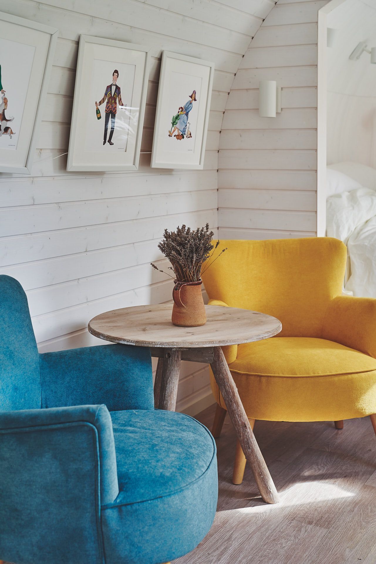 A room with a blue armchair, a yellow armchair, and a small round table with a vase of dried flowers.