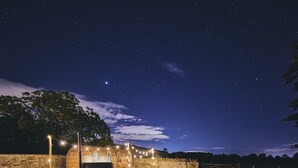 Un'ambientazione rustica all'aperto, con un muro in pietra, una struttura in legno con finestre ad arco e luci a catena.