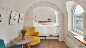 Una camera da letto accogliente con soffitto curvo, un letto con biancheria bianca, una poltrona gialla, un tavolino di legno con un vaso e una finestra con vista sull'esterno.