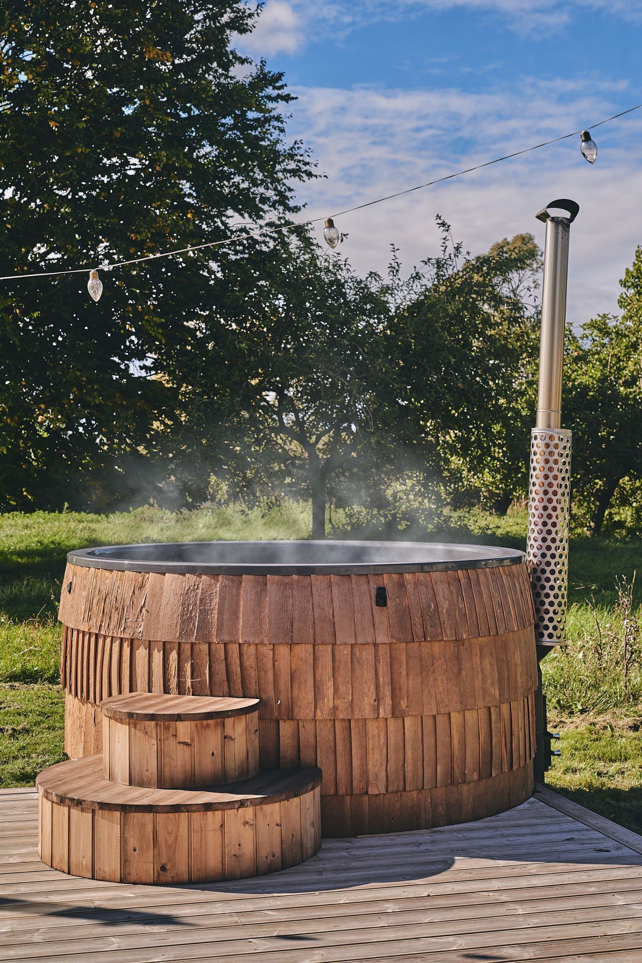 A wooden hot tub with a metal chimney, surrounded by a wooden deck and trees.