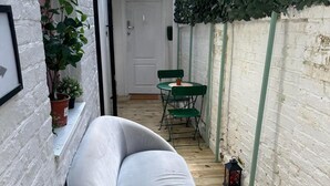 A narrow outdoor space with a white cushioned bench, a small round table with two chairs, and a lantern on the floor. - Studio Sleeps 3 - Outdoor Space - Pet Friendly (London)