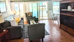 Sala de estar com sofá, poltrona e mesa de centro. Há uma porta de vidro deslizante que dá para uma varanda com vista para a praia.