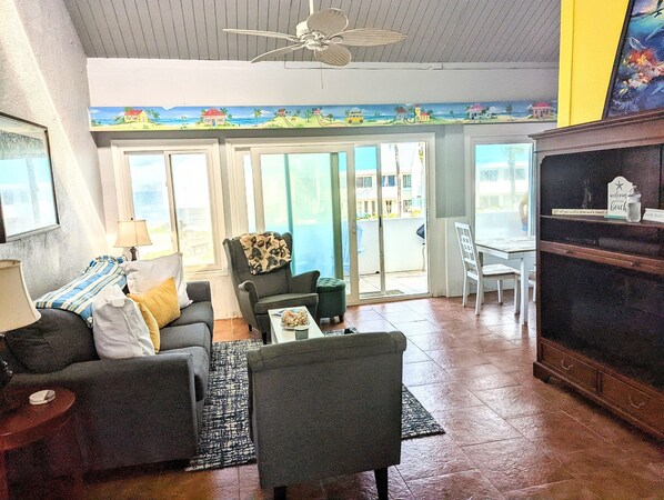 Ett vardagsrum med en soffa, en fåtölj och ett soffbord. Det finns en skjutdörr i glas som leder ut till en balkong med utsikt över stranden.