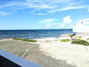 En strandutsikt med en vit byggnad, en stenig kustlinje och en klar himmel.