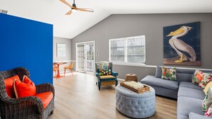 Un salon spacieux avec un plafond haut, un ventilateur de plafond, un coin repas avec des chaises orange et un grand canapé gris.