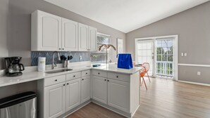 Une cuisine moderne avec des meubles blancs, une machine à café et un évier. Il y a une fenêtre avec des stores et une porte qui donne sur un balcon.