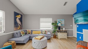 Un salon moderne avec un canapé d’angle gris, un pouf rond, une petite table avec une lampe et un téléviseur fixé au mur.