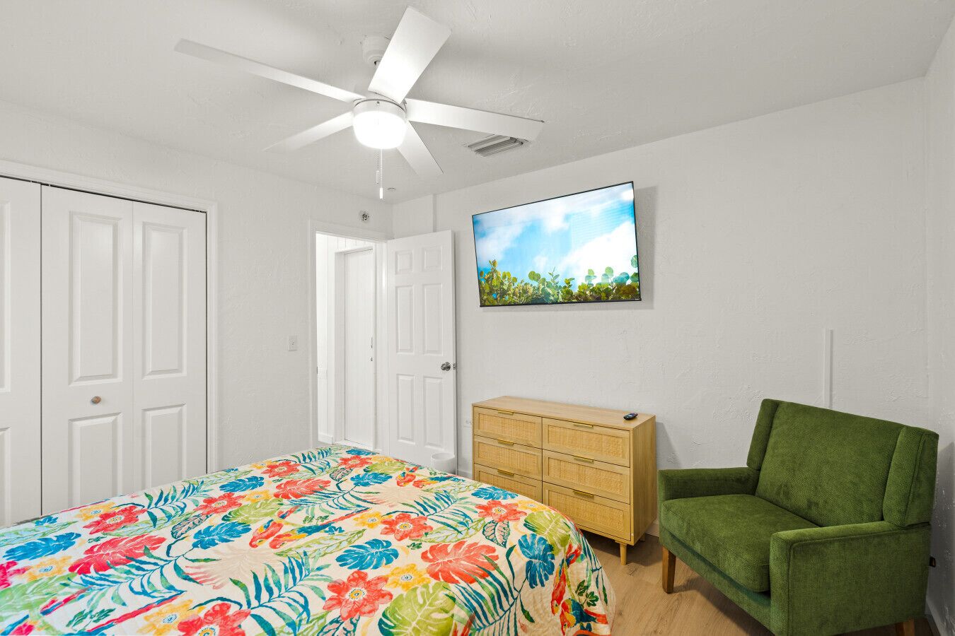 A bedroom with a bed, a green armchair, a wooden dresser, a ceiling fan, and a wall-mounted TV.