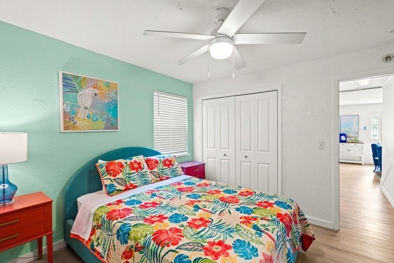 Un dormitorio con pared verde azulado, ventilador de techo, cama con colcha estampada, mesita de noche roja y ventana con persianas.