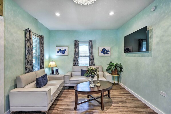 A living room with a sofa, a coffee table, and two windows with curtains. There are framed pictures on the wall and a plant on a side table. - 1 Mi to UTA: Arlington Elm Cottage w/ Fenced Yard (Arlington)