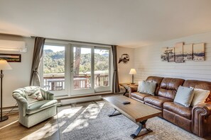 Een woonkamer met uitzicht op een bergachtig landschap, een bruine leren bank, een lichtkleurige fauteuil, een houten salontafel en een open haard.