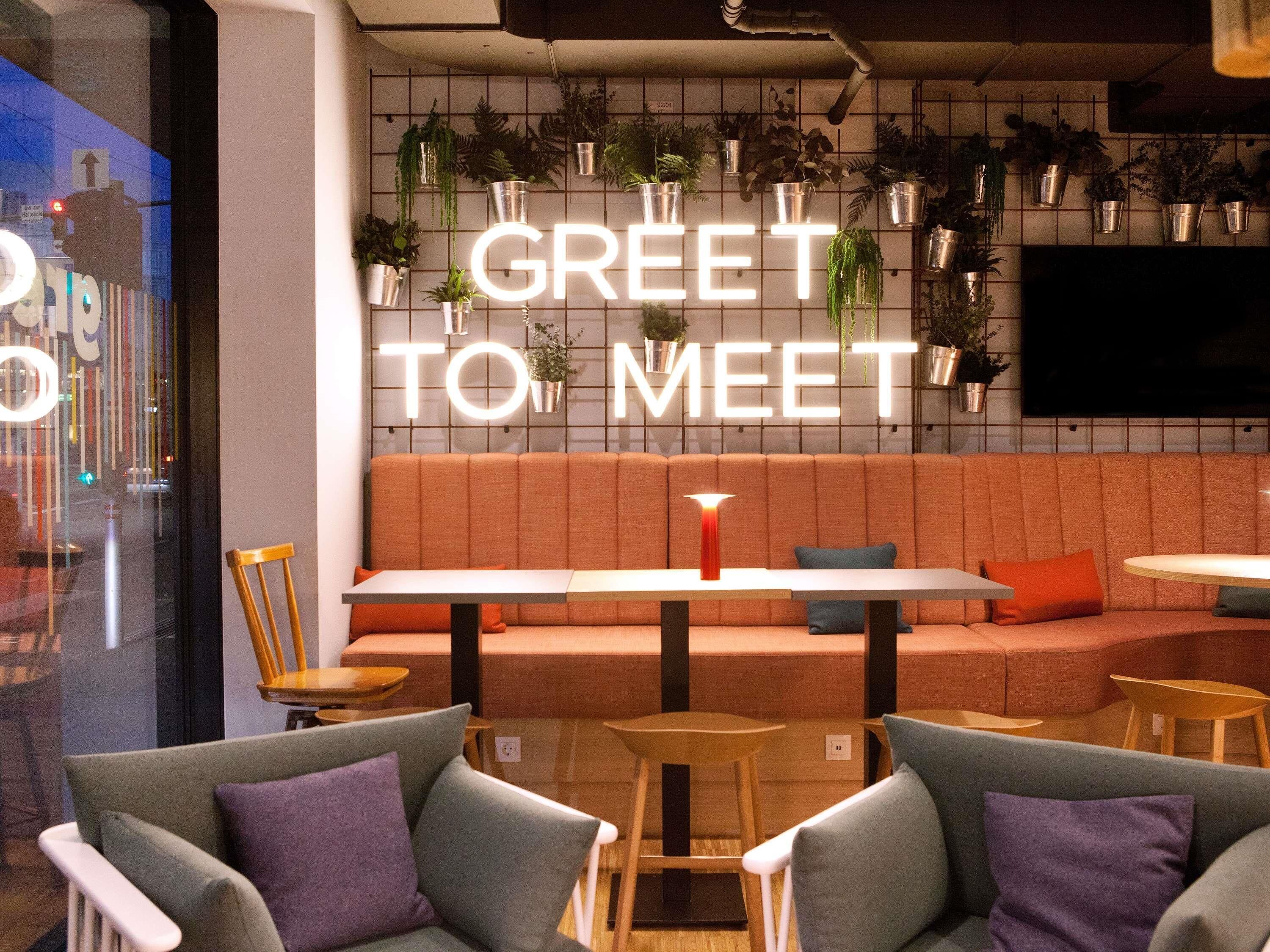 Ein moderner Lounge-Bereich mit einer Couch und Sesseln, einer Neon-Schriftzugtafel mit der Aufschrift "GREET TO MEET" und Topfpflanzen an der Wand.
