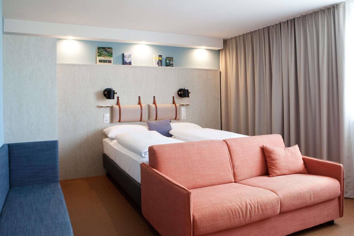 Ein Hotelzimmer mit einem Bett, einem blauen Sessel, einem rosafarbenen Sofa, einem an der Wand montierten Bücherregal und einem Fenster mit Vorhängen.