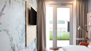 A hotel room with a flat-screen TV mounted on a textured wall, a bed, a desk with a lamp, and a sliding glass door leading to an outdoor area. - Greet Wien City Nord  (Vienna)