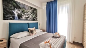 Une chambre d’hôtel avec un lit, une table de chevet, un tableau représentant une cascade et une fenêtre avec des rideaux bleus.