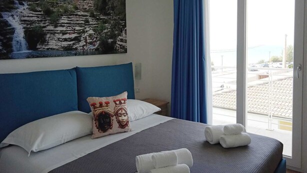 Une chambre d’hôtel avec un lit, un tableau représentant une cascade, une vue sur la mer et un balcon avec des rideaux bleus.
