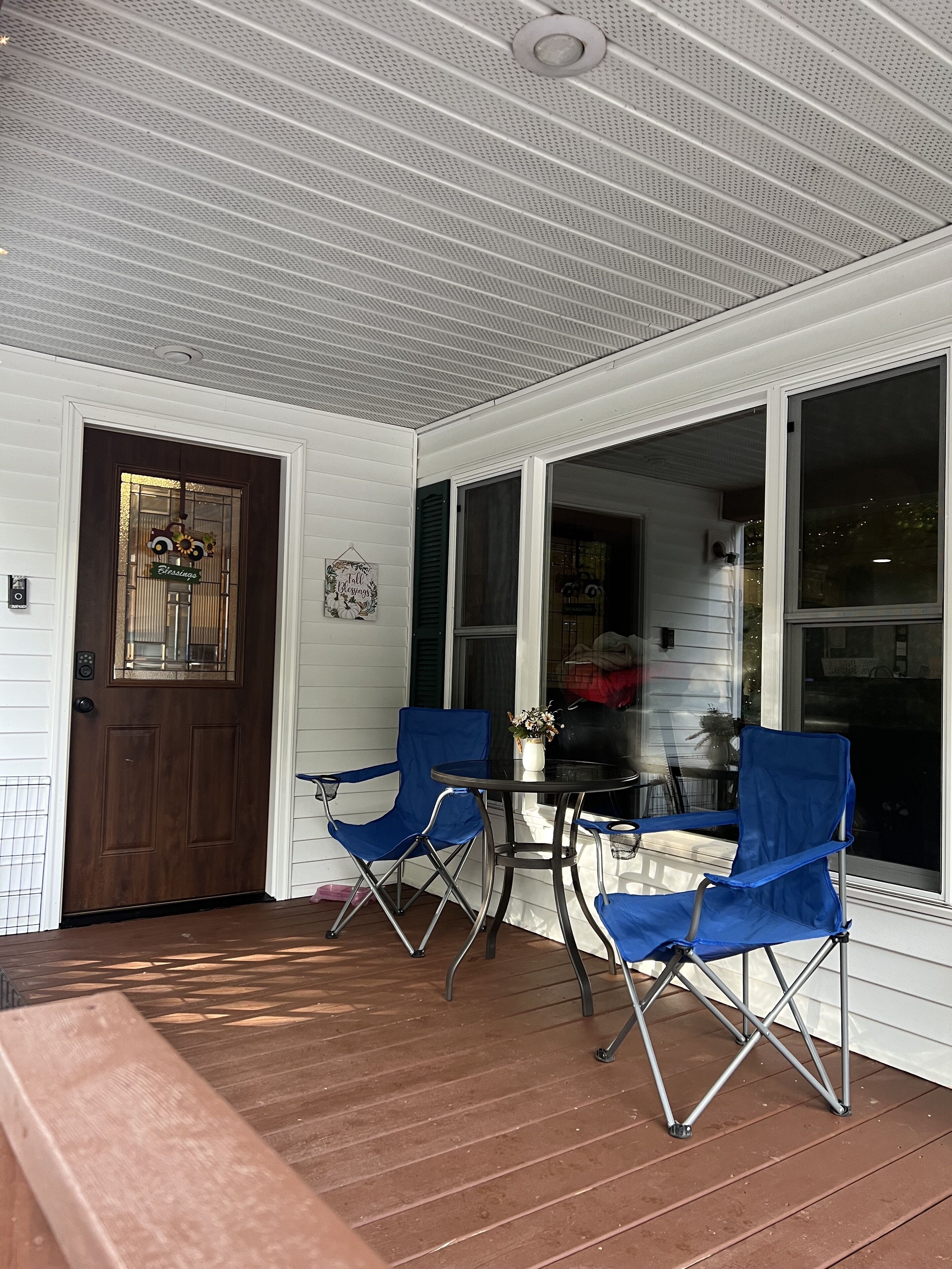 A covered porch with a wooden bench, two blue folding chairs, a small table with a vase, and a door with a 'Blessings' sign.
