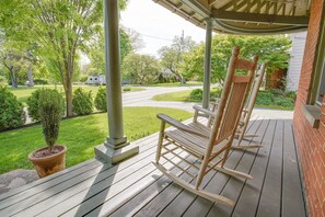 Een houten veranda met een schommelstoel en een bloempot, met uitzicht op een groen gazon en een weg met een huis op de achtergrond.