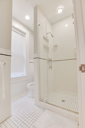 Un baño moderno con ducha de vidrio, azulejos blancos, inodoro y ventana con persianas.