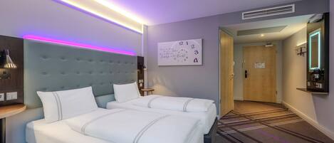 A hotel room with two beds, a headboard, a bedside table, a wall clock, and a carpeted hallway.